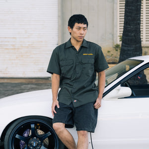 Man in a green shirt standing next to a white car with a palm tree in the background